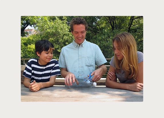 A man shows two learners how to cut a plastic bottle with scissors.