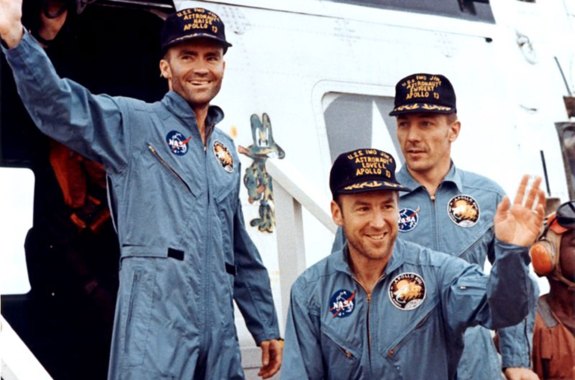 Three men in blue space suits wave outside of an aircraft.