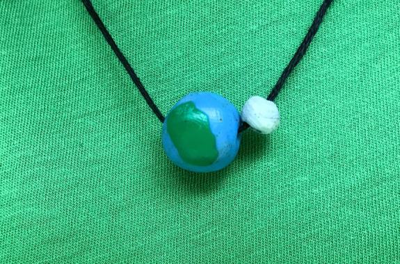 Two handmade clay beads strung on a black nylon string set against a green fabric background. One bead looks like the Earth. The other looks like the Moon.