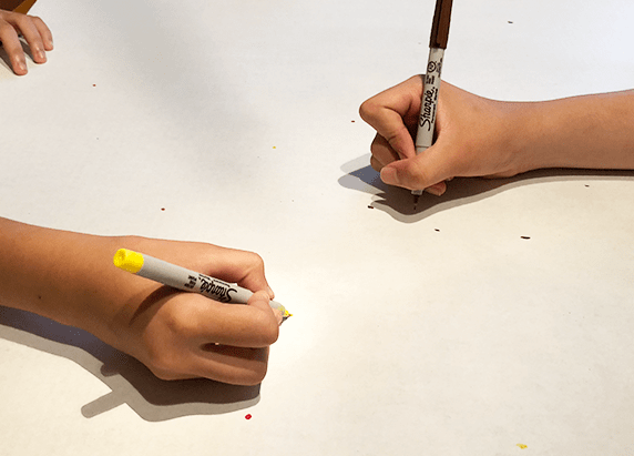 two hands with sharpie pens drawing on a white board