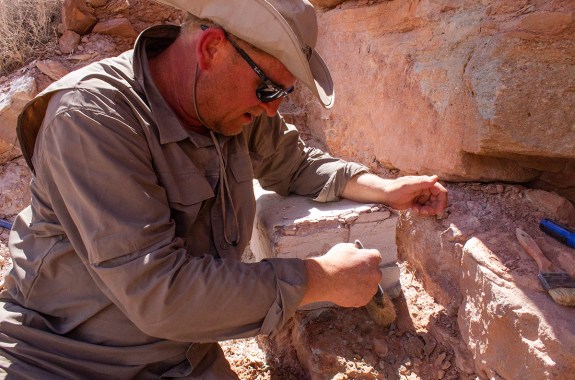 a man sits next to sandstone and brushes away dirt with a brush