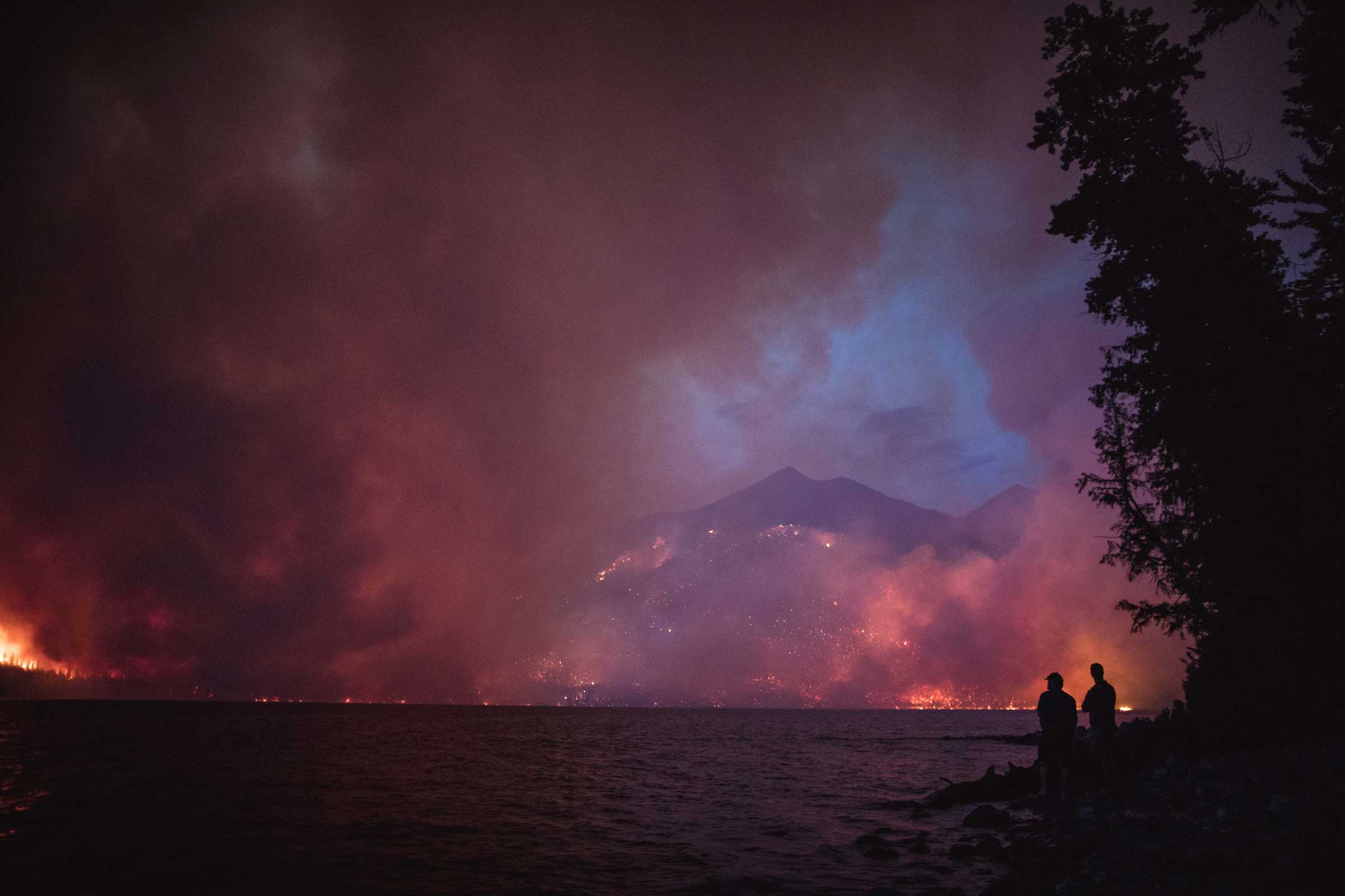 looking across a lake to massive smoke plumes obscuring mountain ranges, taken around dusk