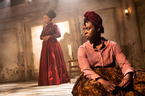 an intense moment with two women of color on the stage. one sits in the foreground looking off into the distance, while the other stands in the background with her hands over her stomach