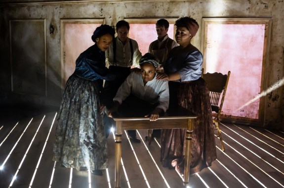 five people around a table on a stage with dramatic lighting shining through slats of the wooden floorboard. two women of color stand on the left of the table assisting another woman of color lying on her belly on the table, while the two men are medically examining her
