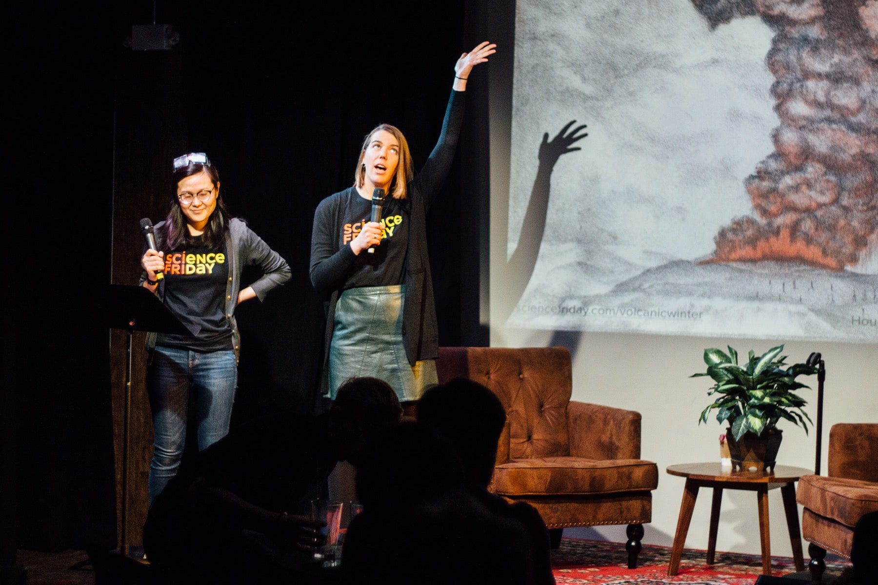 two women on stage with microphones with a painting of an erupting volcano behind them. one of them gestures widely to the sky with her hand