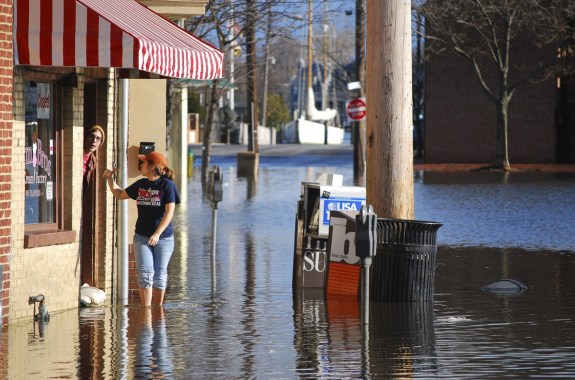 a street is flooded up to a woman's shin. she has her jeans rolled up and is waving to a man who is sticking his head out a door that is barricaded with sandbags.