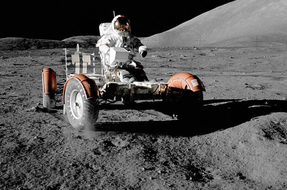 person in space suit riding a vehicle across the rocky gray surface of the moon against black sky