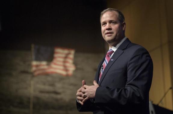 white man with blond hair standing with hands together in front of an image of the moon with a flag in front of it