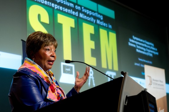 an older black woman in a brightly colored scarf and blue jacket stands at a podium giving a speech. behind her on a projection screen is "symposium on supporting underrepresented minority males in stem"