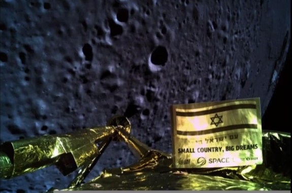 at the bottom of the image is the golden top of the beresheet lunar lander. in the background is the looming, cratered lunar surface. in the foreground of the camera, mounted on the lander, is a small golden sign that features the israeli flag and the words "Small Country Big Dreams" in both English and Hebrew