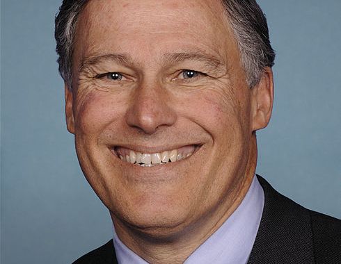 close up portrait of jay inslee grinning against blue background