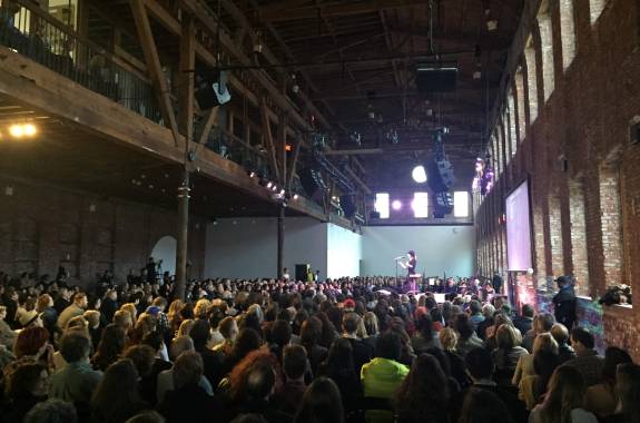 a woman on stage with a mic in a large industrial auditorium space with scores of people watching her