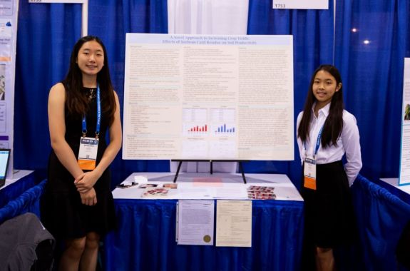 two asian students in front of a science board project. the student on the left is wearing a black dress and the student on the right is wearing a white shirt and skirt