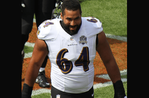 large football player in ravens jersey with number 64 smiling and walking toward camera on black background
