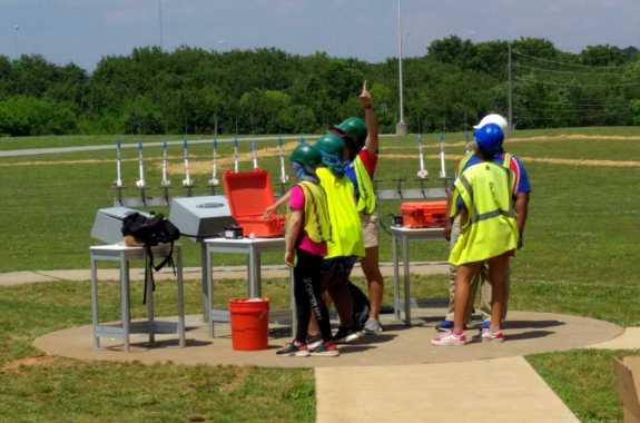 five kids in hard hats and safety vests stand in a grassy field with a row of model rockets