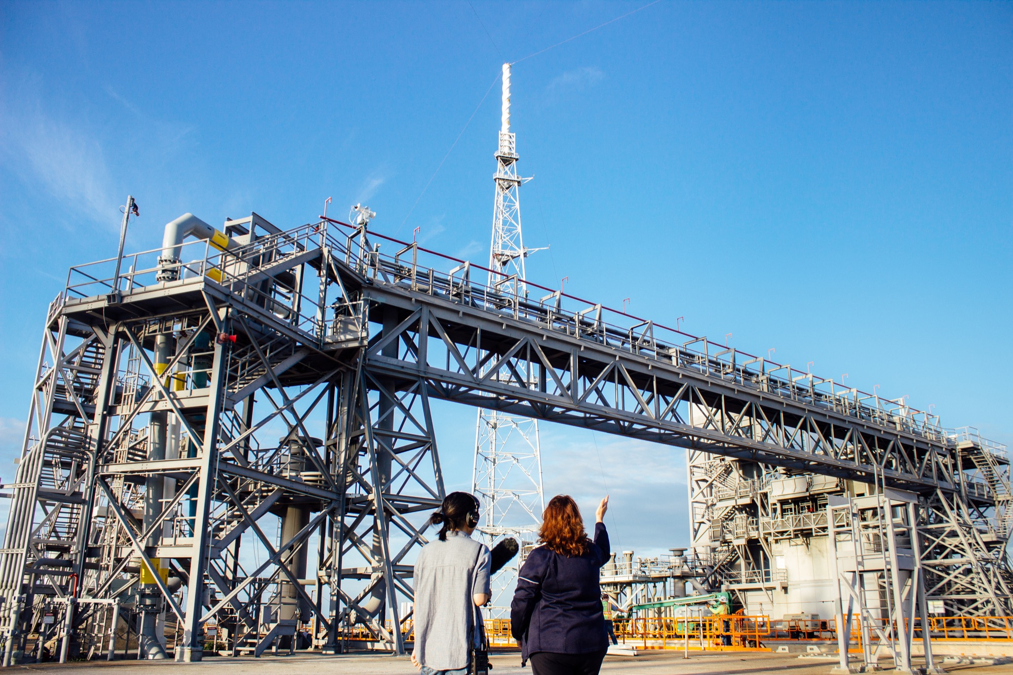 two women stand in front of a giant metal structure, while one of them waves at it with her hand