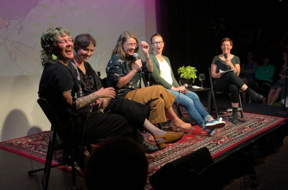 five women stage all with mics laughing with an illustrated bird background behind them