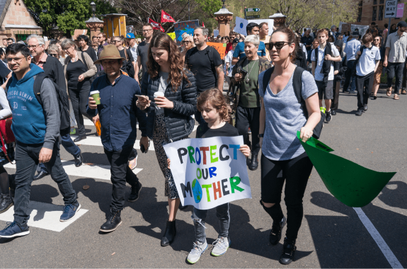 a group of people walking in protest in a street. centered is a woman and child, the child is holding a sign "protect our mother" with the "o" representing the Earth
