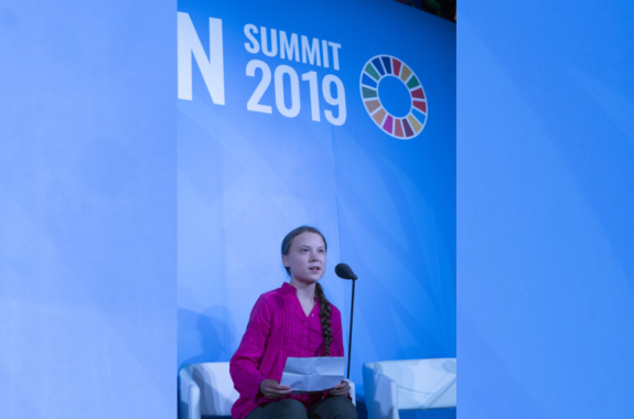 young girl with long braid wearing a reddish-pink shirt sits in front of a microphone giving a speech, with UN Climate branding in the background