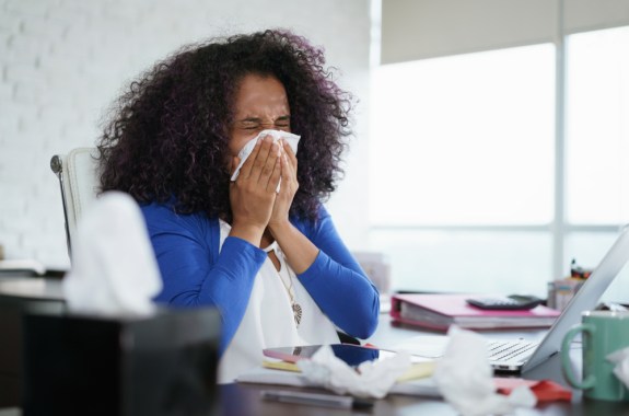 Ill young black woman, sitting at desk with laptop computer and sneezing.