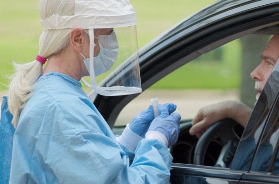 a doctor in protective equipment and mask holding a vial next to a car driver window talking with a man in the car