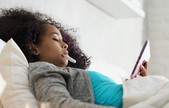 Sick girl with flu lying in bed at home, checking temperature with thermometer and on her tablet
