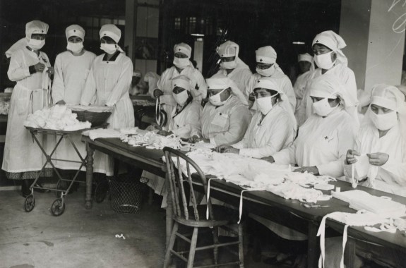 medical workers in face masks and personal protective garb sit at a long table making masks