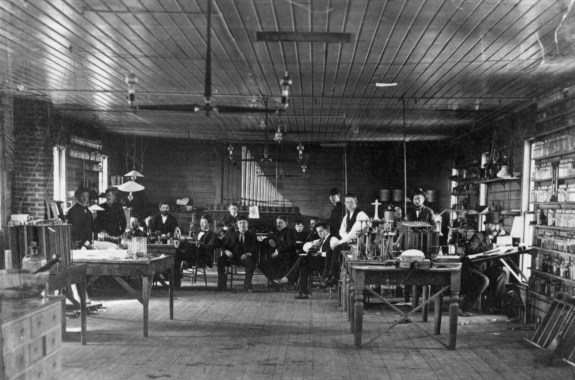 a black and white photo of a group of over a dozen people in a room with lightbulbs and chairs and tables