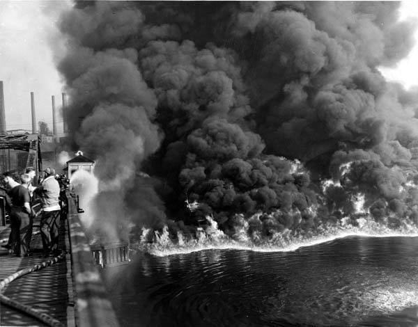 large black clouds of smoke tower above a river on fire. people stand on a bridge with water hoses trying to control the fire