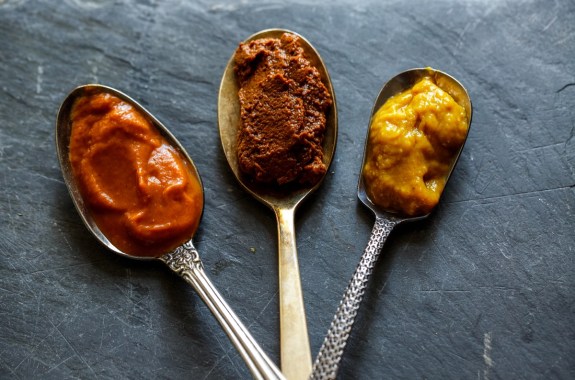 three pastes on spoons, one is a orange-red, the middle is brown and grainy, the third is a yellow-mustard color