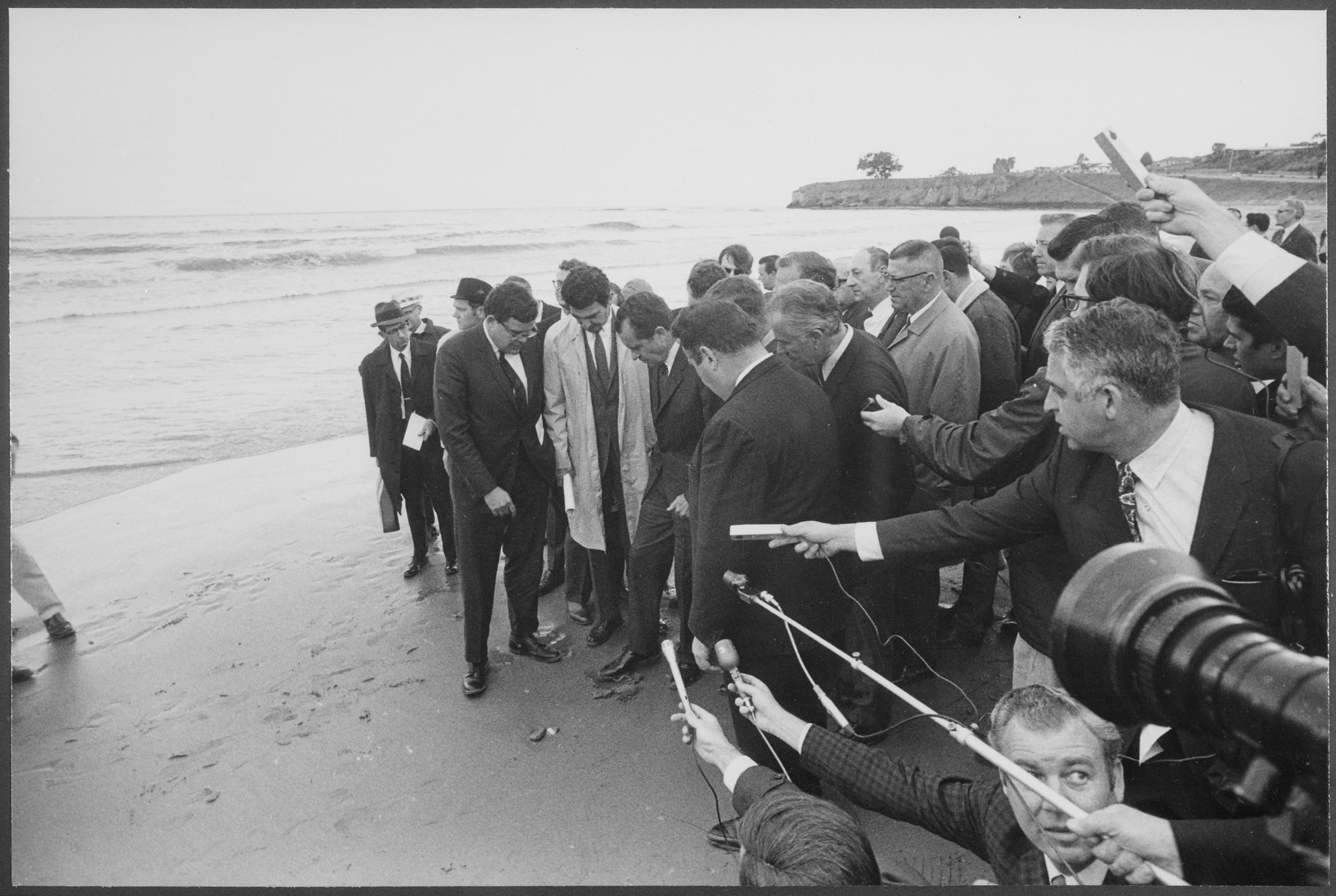 a group of reporters follow president nixon on a beach as they press their feet into the oil covered sand