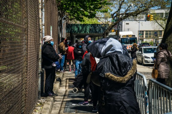 people of color wearing face masks line up outside of a building along a wire fence
