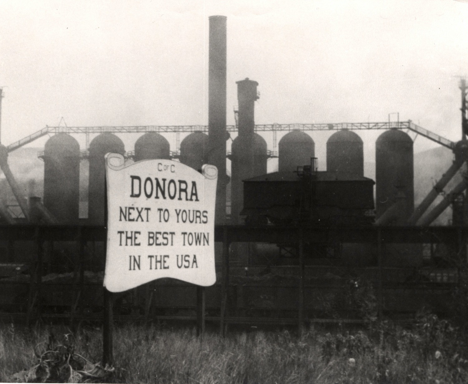 a smoggy factory. in front of it is a sign that reads "donora next to yours the best town in the usa"