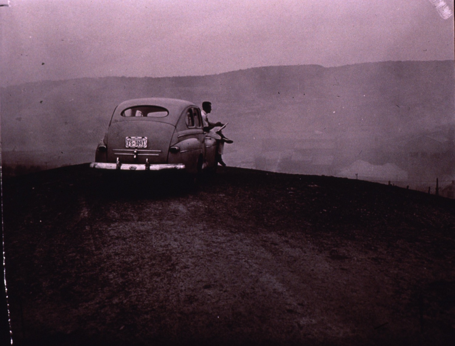 a person sits on top of an old car looking out over a hill at a smoggy sky