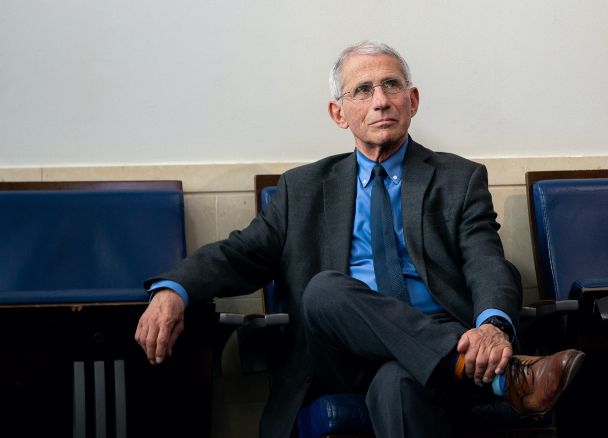 a white man with glasses and wearing a suit sits beneath a blue oval sign of the white house
