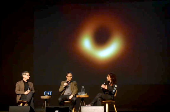 two men and a woman sitting on a stage and speaking into microphones. in the black is an orange ring - the light from the event horizon of a black hole