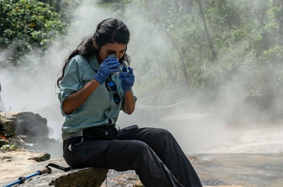a woman scientist wearing gloves looks a specimen through a microscope as she sits on rocks along a steaming river