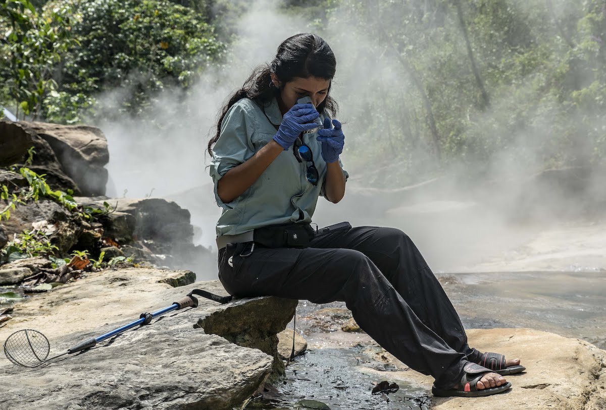 a woman scientist wearing gloves looks a specimen through a microscope as she sits on rocks along a steaming river