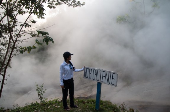 a woman in a hat stands next to a sign that reads "agua caliente" with huge clouds of steam behind her