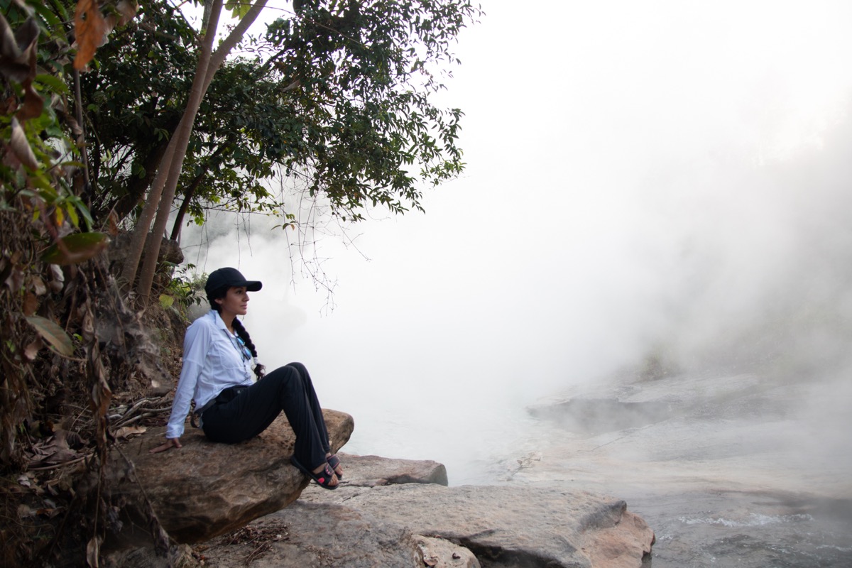 a woman in hat sits next to a river, there is so much steam coming off the river it looks like she is sitting in a cloud