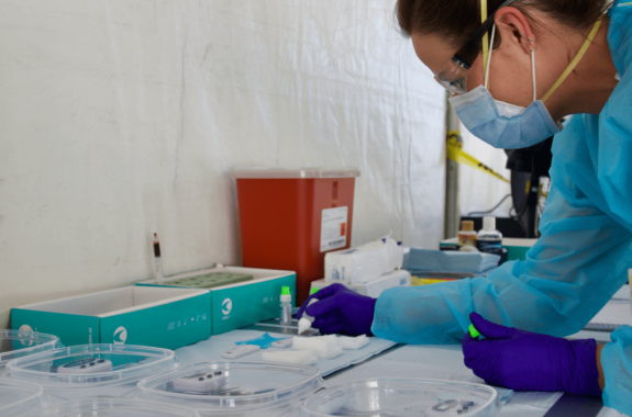 a nurse in a makeshift tent wearing protective gear readies multiple test kits in clear plastic containers
