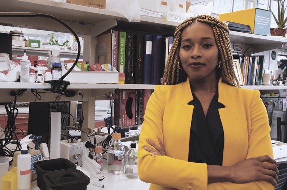 A black woman, Bianca Jones Marlin, stands in a yellow blazer with her arms crossed, in a crowded lab office with lots of items on shelves behind her.
