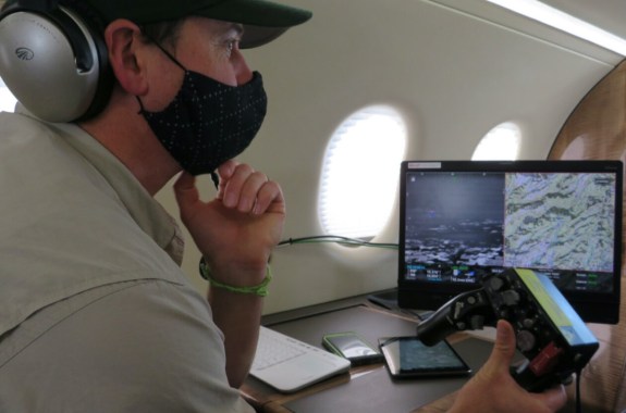 a man wearing a face mask holds a video game-like remote control and looks at a computer screen of maps. he's inside of a plane cabin