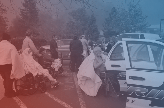 senior adults in wheelchairs and draped with blankets as they are escorted by police cars and rescue teams after a fire. in the background are burned trees and an orange haze. the image has a red and blue filter