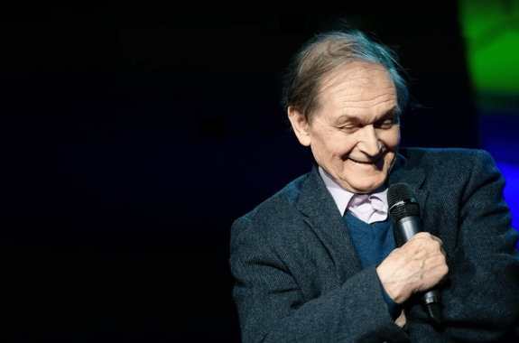 a White man on stage laughing holding a microphone