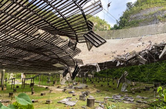 the underside of a massive dish structure, where metal panels have fallen off and some are hanging from the dish