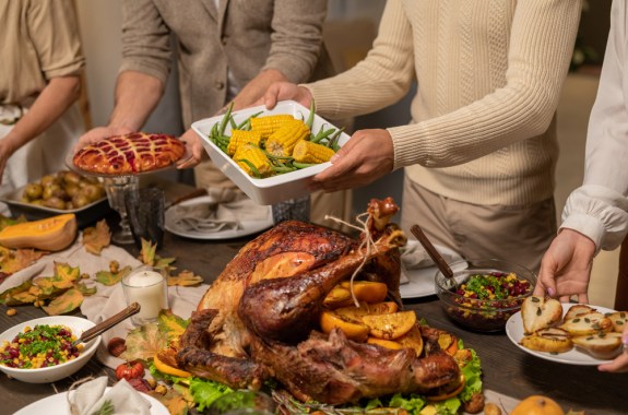 Four members of family putting plates and bowls with homemade dessert, baked corn, salad, sweet pie and other food on table before celebration