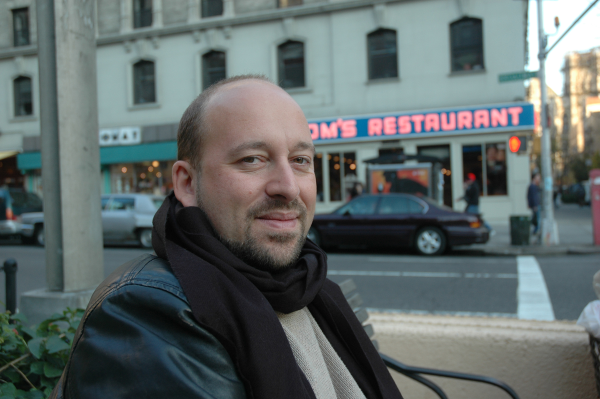 a White man smiling at the camera on the street with tom's restaurant, the restaurant from seinfeld, in the background