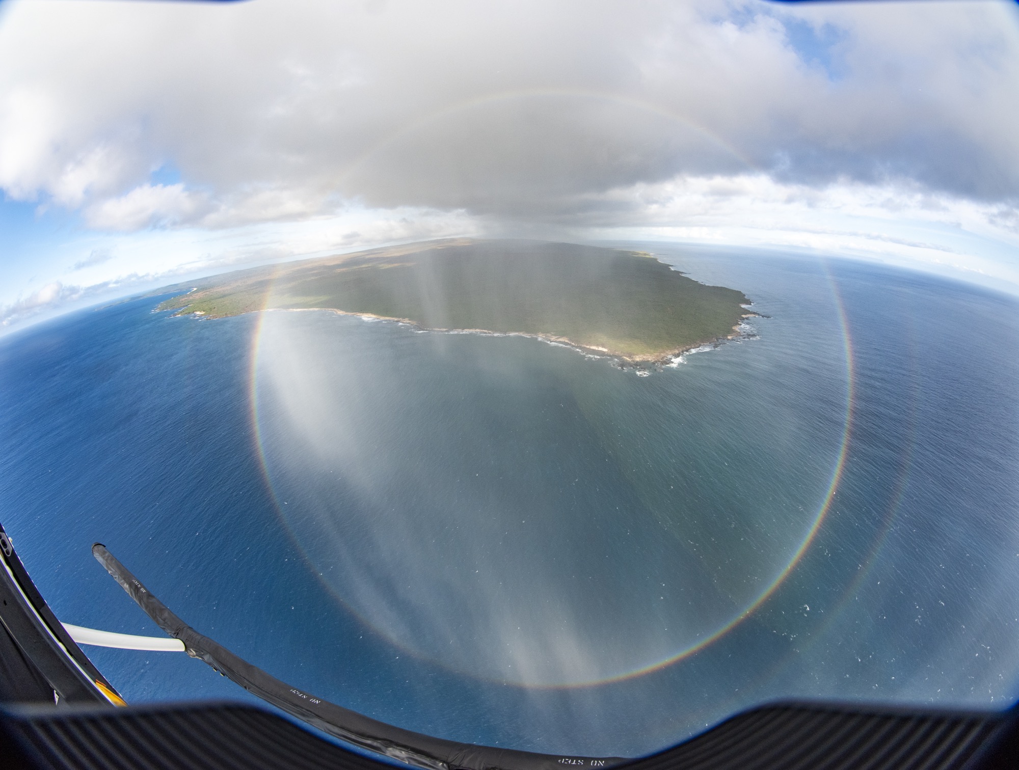 an aerial view of a perfect circular rainbow, taken from a helicopter. in the background you can see an island and vast blue ocean