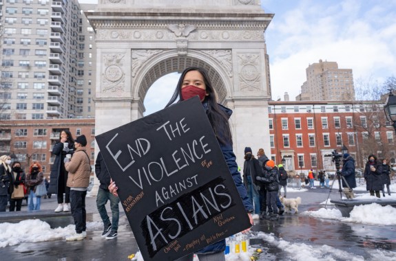 a young asian woman holds a sign that reads end the violence against asians in a snow covered plaza with an archway in the background